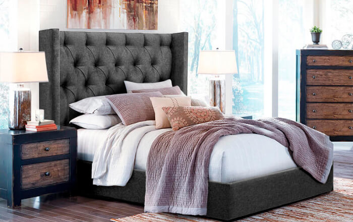 Bedroom with grey bed and headboard with white and light pink bedding complemented by black  metal and wooden dresser and pedestal